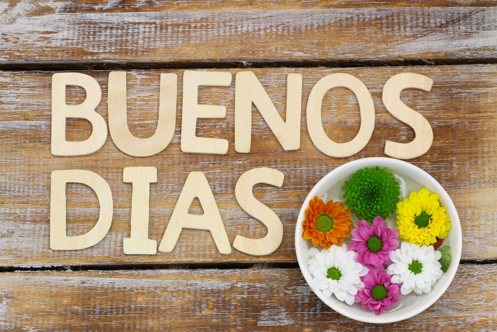 Photo of multicolored daisy flowers in a cup on a wooden table with the letters "BUENOS DIAS".